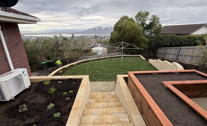 Sandstone-block retaining walls and steps terracing a backyard, with timber raised beds and views over Hobart