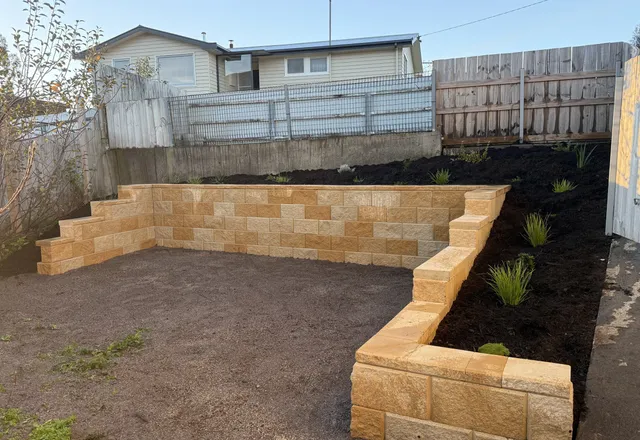 Sandstone-block retaining wall wrapping a levelled backyard, with a freshly mulched and planted bank above